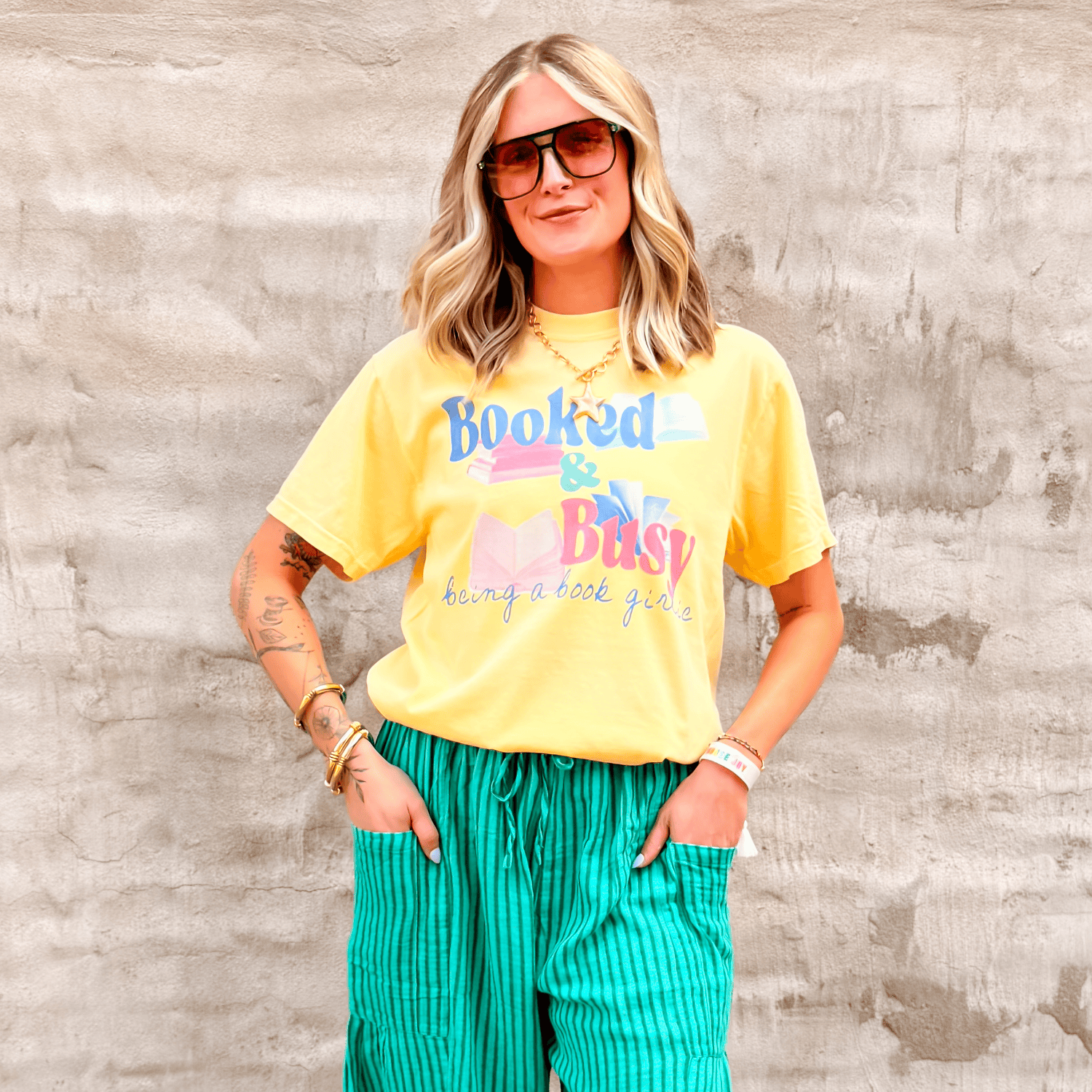 Yellow Booked & Busy tee with colorful text, worn by a woman with sunglasses and green striped pants against a textured wall.