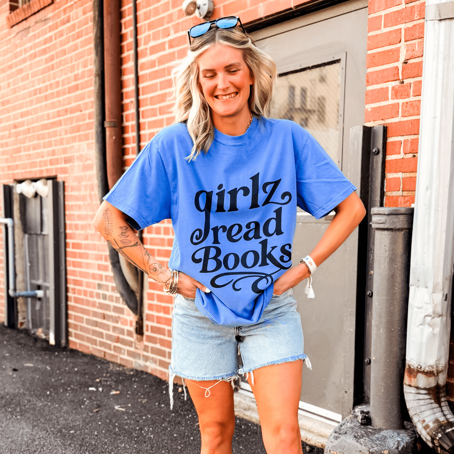Blue Girlz Read Books tee worn by a smiling woman, paired with denim shorts, standing against a brick wall.