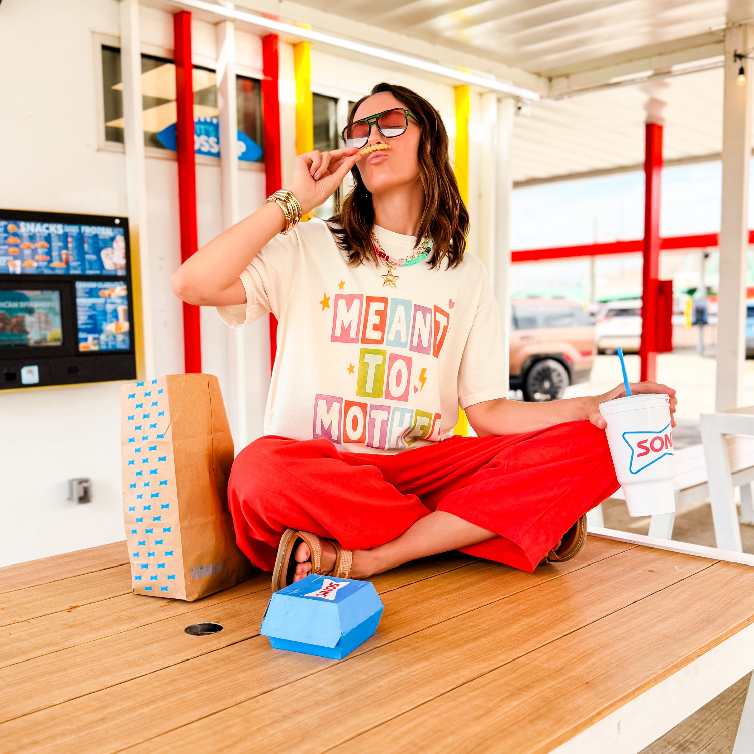 Meant To Mother tee in cream, worn by a woman sitting cross-legged with red pants, enjoying a drink and snacks.