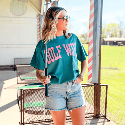 Woman wearing a teal 'GOLF WIFE' shirt holding a golf club outdoors.
