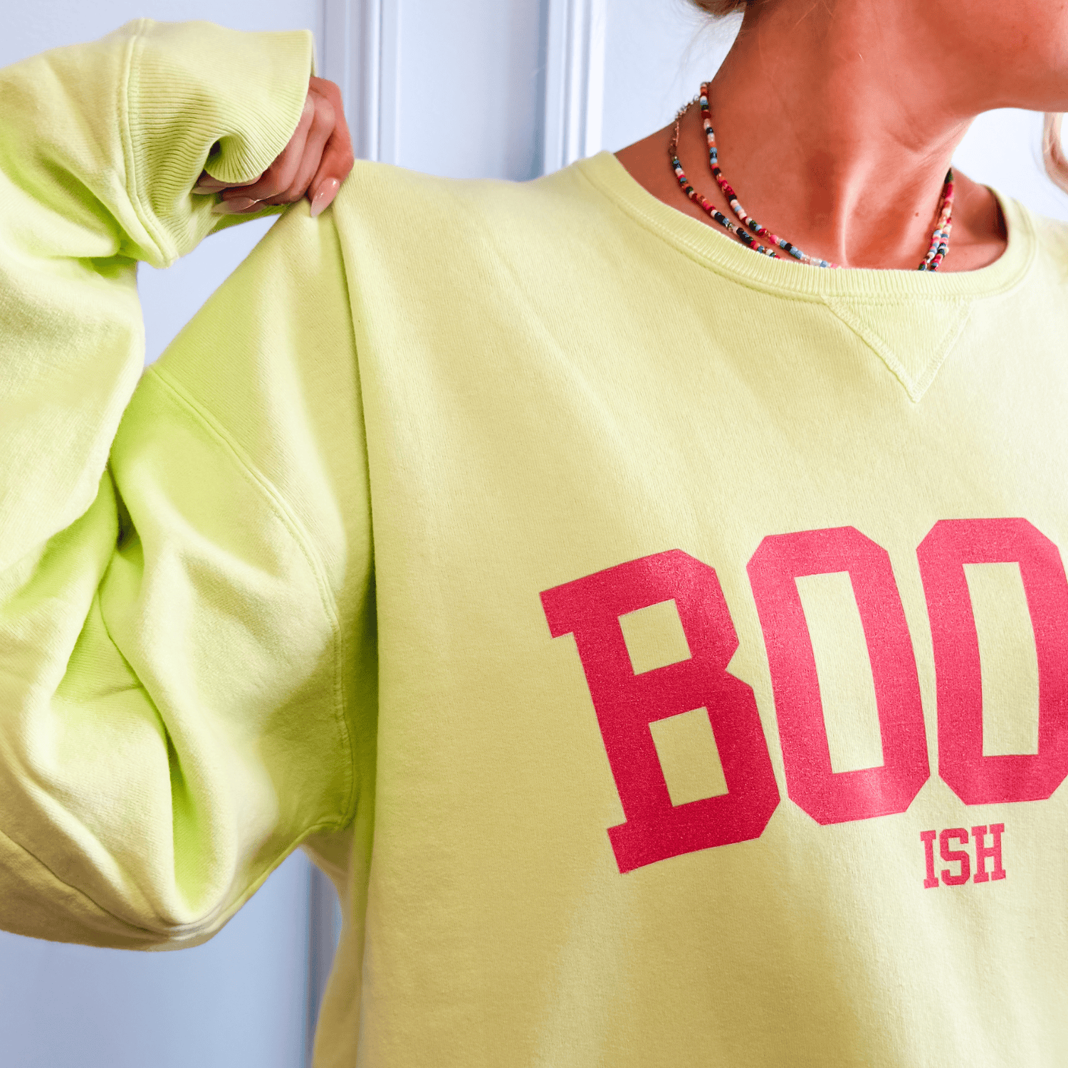 Bookish Comfort Wash crewneck in light yellow with bold red "BOOKISH" text on the front, worn by a person.
