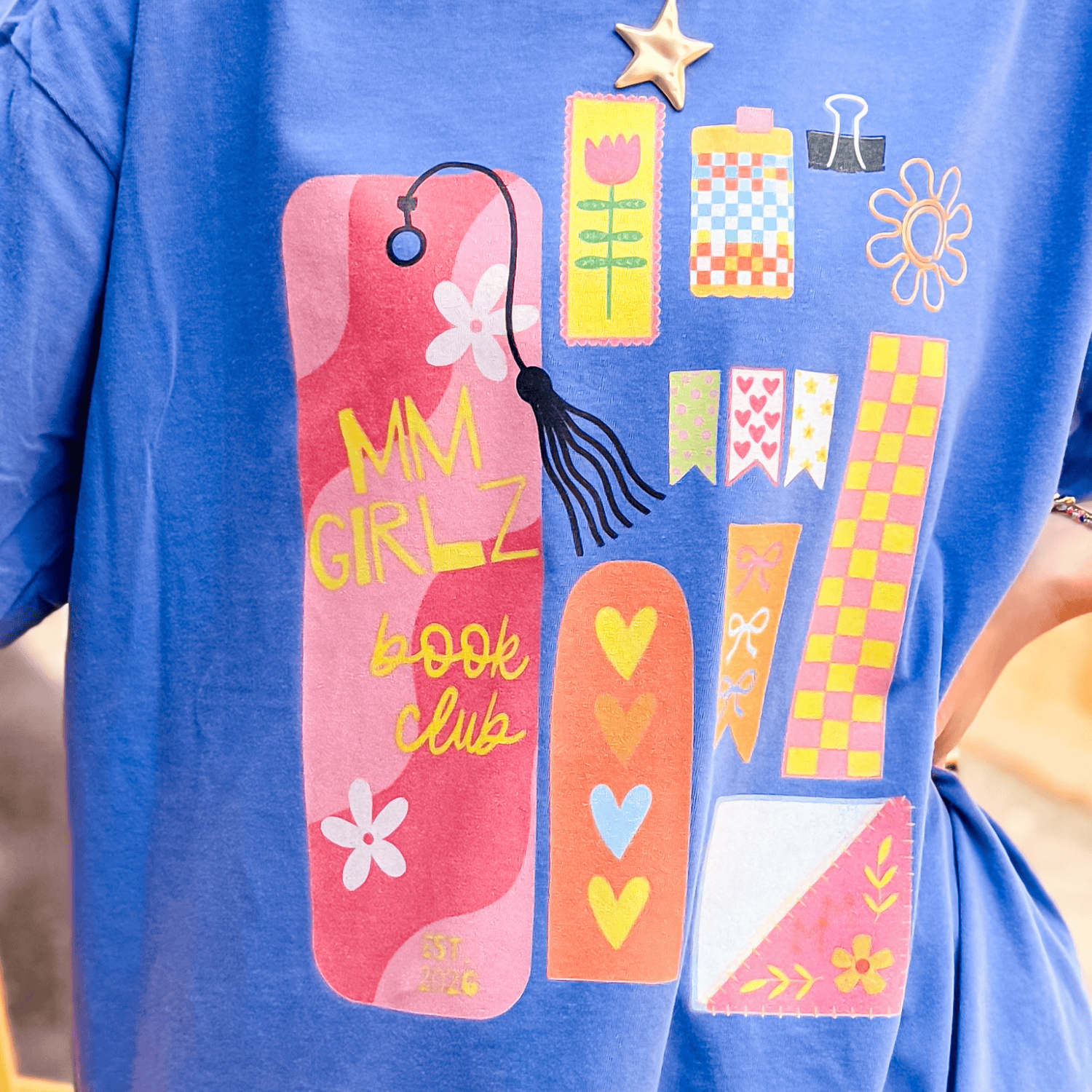 Blue Book Club Tee featuring colorful graphic designs, including flowers, hearts, and a tassel bookmark.