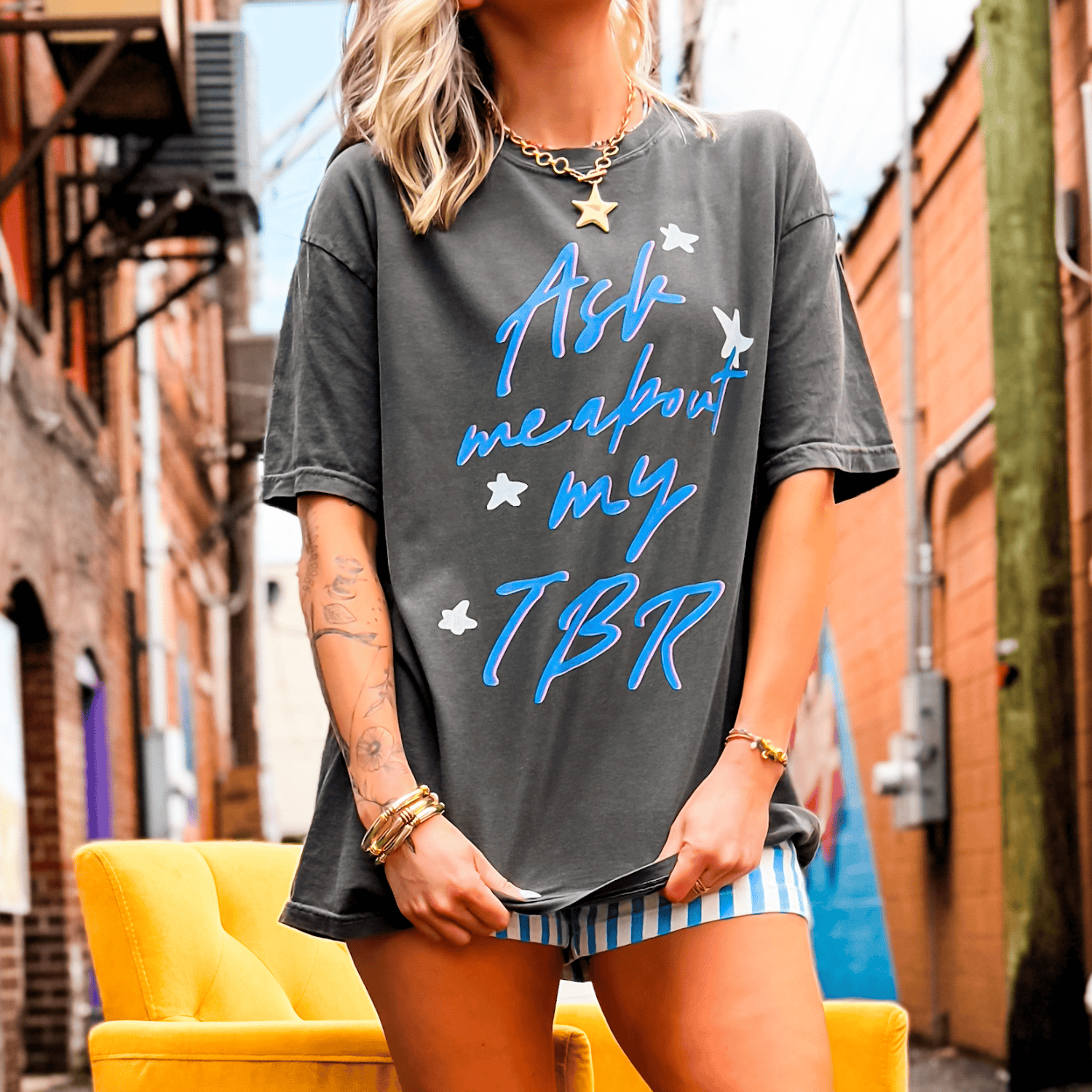 Gray oversized tee with "Ask me about my TBR" in blue, worn by a model in a colorful urban setting.