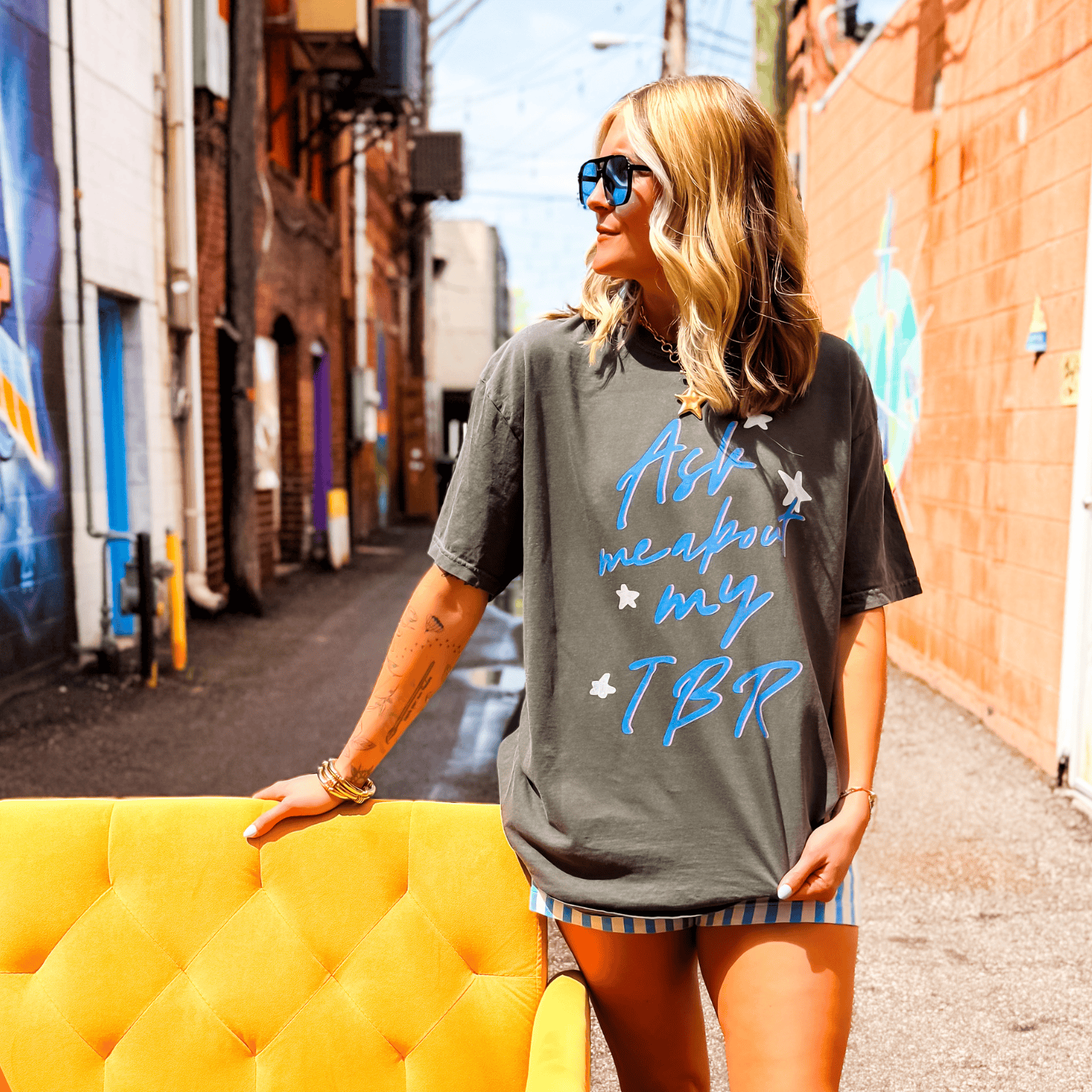 Gray oversized tee with "Ask me about my TBR" in blue script, worn by a woman in sunglasses, standing beside a yellow couch.