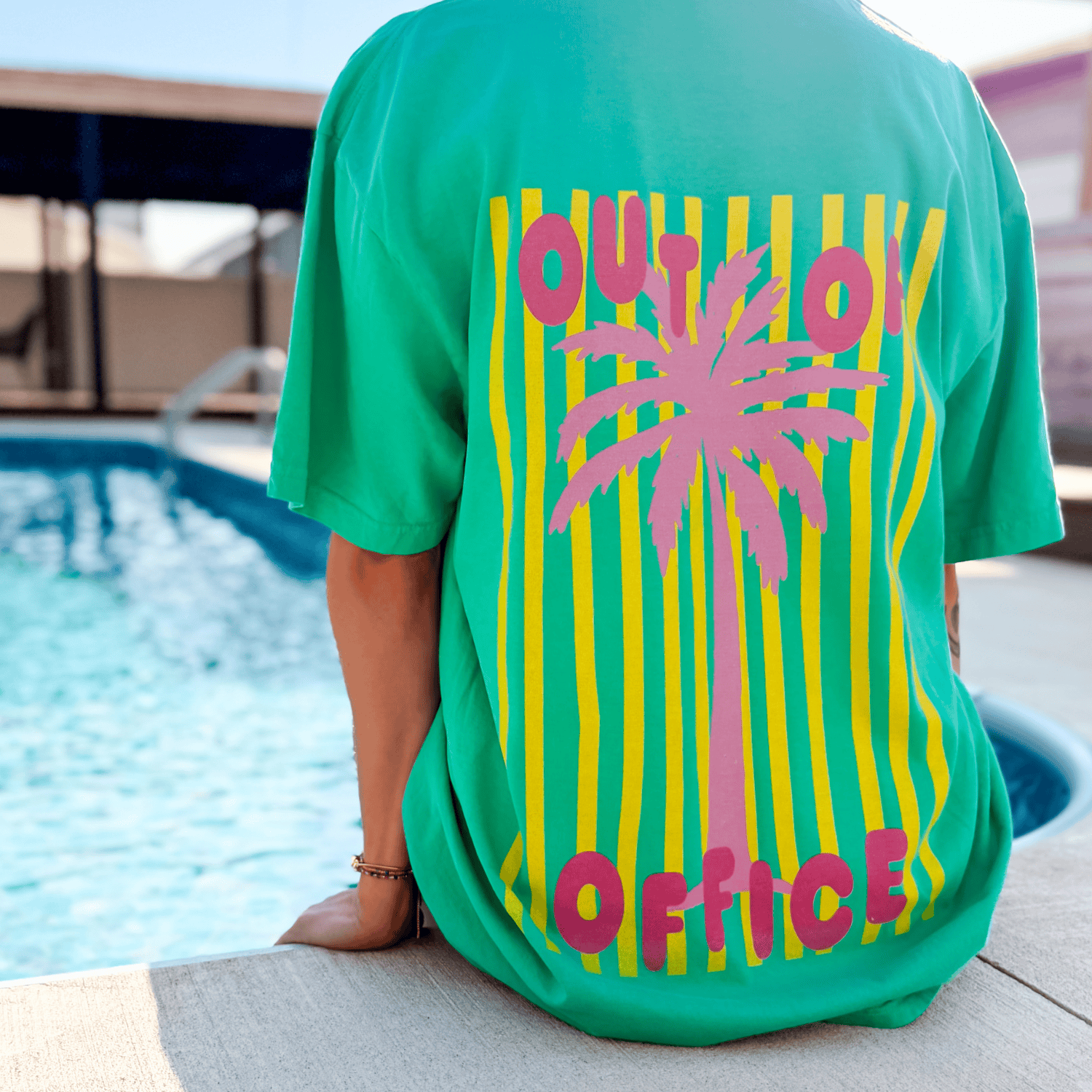 Neon Out of Office tee in green with a pink palm tree graphic and yellow stripes, worn by a person near a pool.